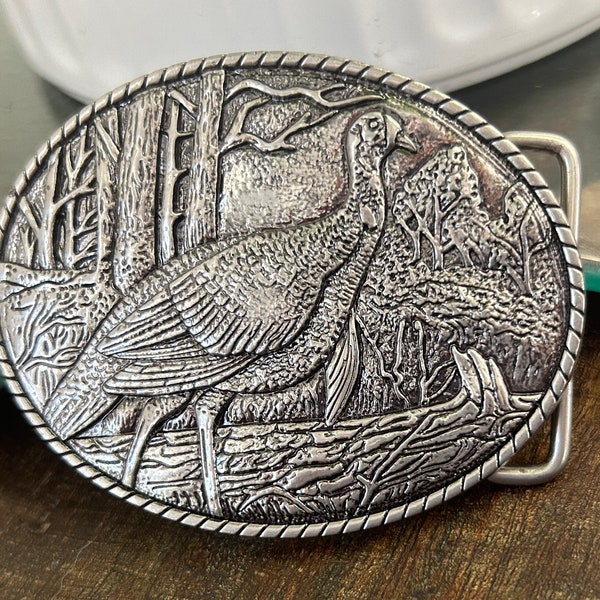 Puede incluir: Hebilla de cinturón de plata con un diseño detallado de un pavo salvaje en un entorno de bosque.