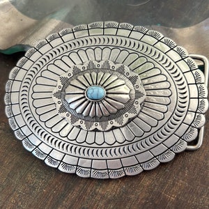 May include: A silver belt buckle featuring a turquoise stone at its centre. The buckle has a circular design with elaborate details.