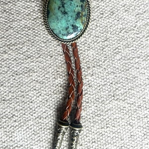 May include: A bolo tie featuring a large oval turquoise stone set in a silver-toned metal frame. The braided leather cord is brown, with decorative silver-toned metal tips.