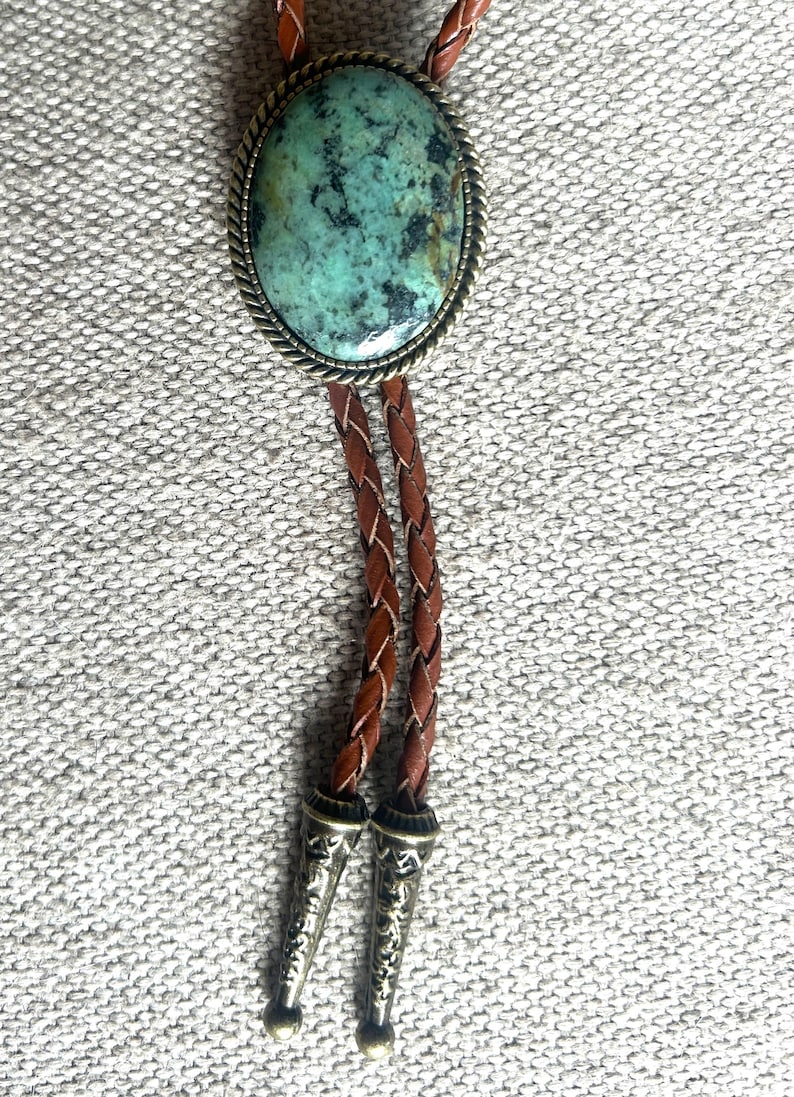May include: A bolo tie featuring a large oval turquoise stone set in a silver-toned metal frame. The braided leather cord is brown, with decorative silver-toned metal tips.