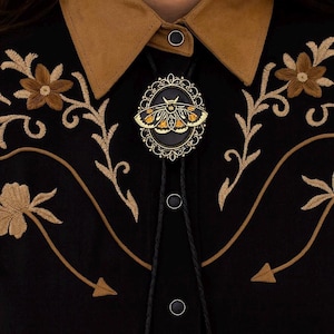 Puede incluir: Camisa negra con cuello marrón y bordado floral. Una corbata bolo con un colgante ovalado decorativo con un diseño de polilla dorada y naranja es el punto focal. La camisa tiene cierre de botones.