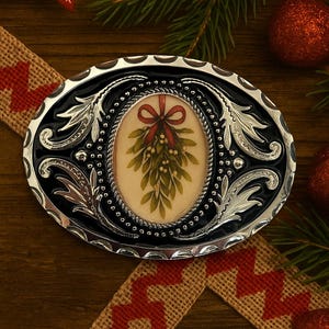 May include: Oval silver and black belt buckle featuring a mistletoe design. The buckle has a black centre with a silver border and decorative flourishes. A red ribbon accents the mistletoe. Christmas decorations surround the buckle.