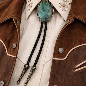 May include: A turquoise bolo tie with a braided black cord and ornate metal tips. The tie is set against a cream-colored shirt and a brown suede jacket with decorative stitching and silver buttons. The collar of the shirt has embroidered floral details.