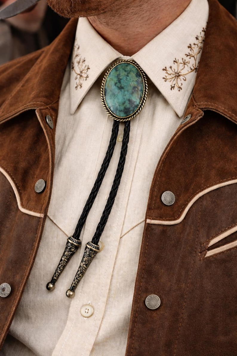 May include: A turquoise bolo tie with a braided black cord and ornate metal tips. The tie is set against a cream-colored shirt and a brown suede jacket with decorative stitching and silver buttons. The collar of the shirt has embroidered floral details.