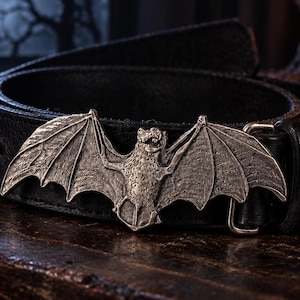 Op de afbeelding: Een zwarte leren riem met een grote, gedetailleerde zilverkleurige metalen gesp in de vorm van een vleermuis met gespreide vleugels. De gesp heeft een getextureerde, antieke afwerking en de riem is gemaakt van glad, donkerzwart materiaal met stiksels.