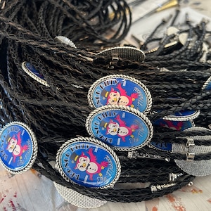 Puede incluir: Un mont&oacute;n de corbatas bolo trenzadas negras con dijes ovalados plateados que presentan una imagen de dibujos animados de dos p&aacute;jaros rojos con un sombrero de copa y un mu&ntilde;eco de nieve. Los dijes tienen fondos azules con texto blanco que dice "The Big Philly 2023 New Year's Eve".