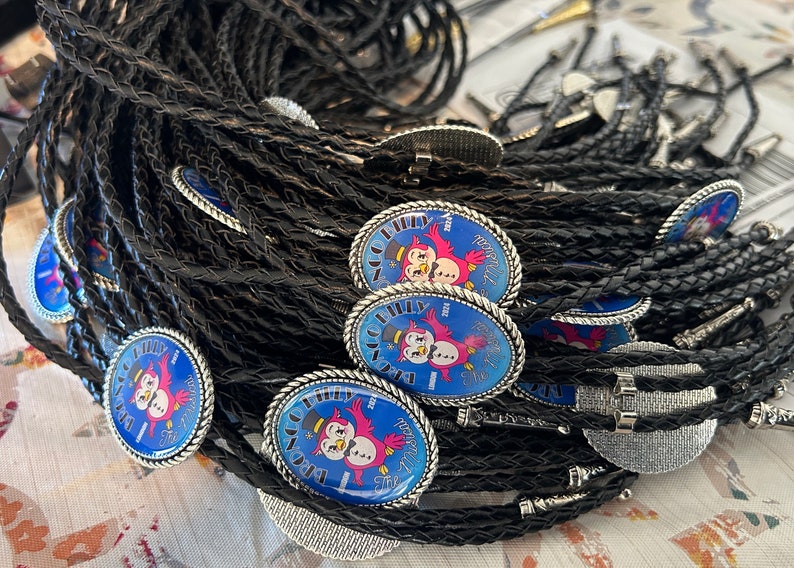 Puede incluir: Un mont&oacute;n de corbatas bolo trenzadas negras con dijes ovalados plateados que presentan una imagen de dibujos animados de dos p&aacute;jaros rojos con un sombrero de copa y un mu&ntilde;eco de nieve. Los dijes tienen fondos azules con texto blanco que dice "The Big Philly 2023 New Year's Eve".