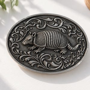 Puede incluir: Una hebilla de cinturón de metal con forma ovalada que presenta una figura de armadillo detallada y en relieve en el centro. El armadillo está rodeado por un intrincado patrón de volutas en relieve que enmarca el borde del accesorio plateado.