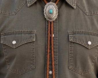 May include: A bolo tie featuring a braided brown leather cord and a silver-toned oval concho centerpiece with a central turquoise stone. The tie ends in two matching silver-toned metal tips with engraved detailing, worn over a dark olive shirt.