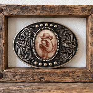 Op de afbeelding: Een ovale metalen riemgesp met een centrale sepia-kleurige illustratie van een wasbeer die een cowboyhoed draagt en mondharmonica speelt. De gesp heeft een donkere, in reliëf gemaakte bloemenrand met zilverkleurige noppen.
