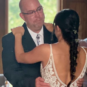 Puede incluir: Una novia con un vestido de novia blanco de encaje con la espalda baja y un peinado trenzado baila con su padre. &Eacute;l lleva un traje negro y una camisa blanca con una corbata bolo.