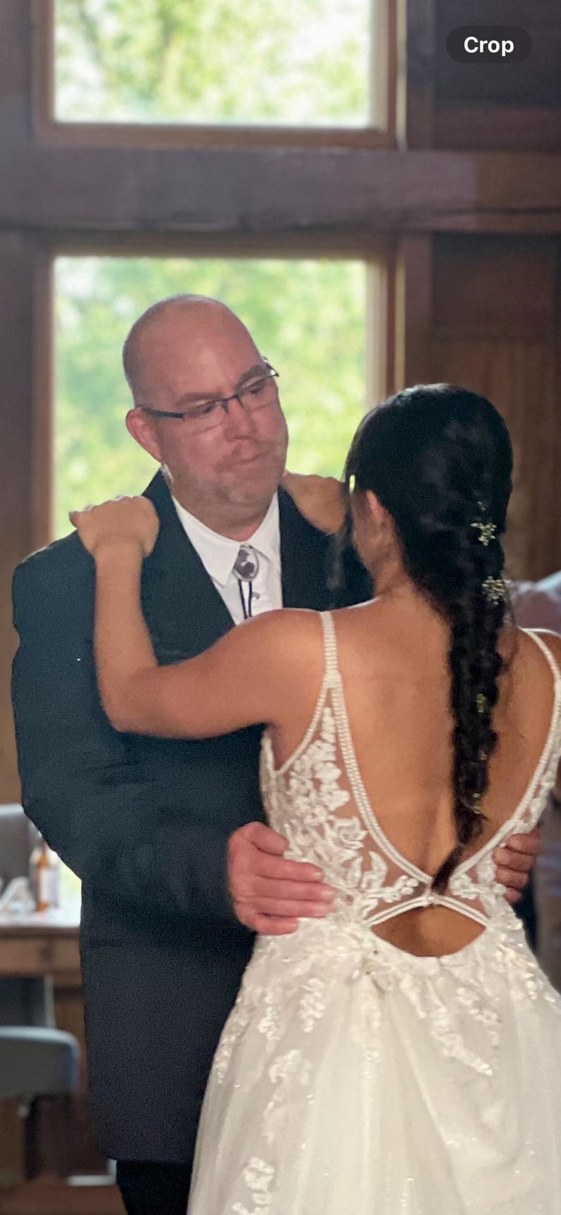 Puede incluir: Una novia con un vestido de novia blanco de encaje con la espalda baja y un peinado trenzado baila con su padre. &Eacute;l lleva un traje negro y una camisa blanca con una corbata bolo.