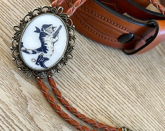 May include: A brown leather bolo tie with a silver-toned oval pendant featuring a cartoon cat holding a gun. The pendant has a white background and intricate details around the edge.