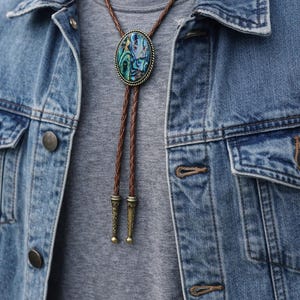 May include: A bolo tie with a brown braided leather cord and decorative metal tips. The focal point is an oval pendant with a colorful, iridescent abalone shell design, set in a bronze-colored metal frame. The tie is worn over a gray shirt and denim jacket.