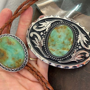 May include: A bolo tie and belt buckle set featuring oval turquoise stones. The bolo tie has a braided leather cord. The buckle has a black background with silver filigree and a silver border. The turquoise stones have brown and green hues.
