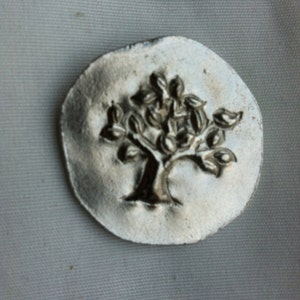 May include: A silver charm with a tree design. The tree is embossed on the charm and has a detailed trunk and branches.