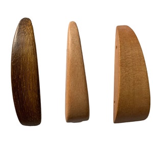 Può includere: Tre ganci per cappotti in legno in diverse tonalità di marrone. I ganci hanno la forma di un triangolo arrotondato e una superficie liscia leggermente testurizzata.