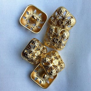 May include: Five gold-colored metal buttons with intricate floral designs. The buttons are square shaped and have a raised, ornate pattern.