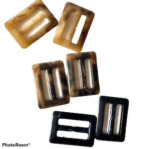 May include: Five plastic belt buckles in various shades of brown and black. The buckles have a rectangular shape with two parallel slots.