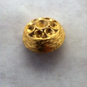 Op de afbeelding: Een gouden metalen knoop met een decoratief ontwerp. De knoop heeft een verhoogd midden met een cirkelvormig patroon en een getextureerde buitenrand.