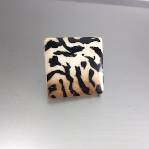 May include: A square, decorative button with a cream-colored background and a black and brown animal print pattern. The button has a glossy finish and is set against a gray background.