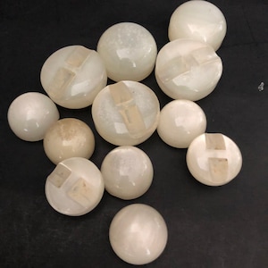 May include: A collection of white, round, plastic buttons with a raised, clear plastic design in the center. Some buttons have a cross-shaped design, while others have a single, vertical line.