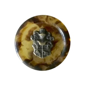 May include: A round, brown and beige button with a silver metal crest in the center. The crest features a stylized design with a crown and other details.