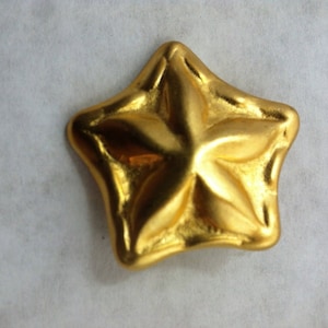 Puede incluir: Un colgante de color dorado en forma de estrella de cinco puntas con una superficie texturizada.