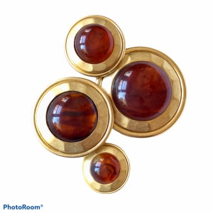 May include: A gold tone brooch with four round, amber-colored stones set in gold tone bezels. The stones are of varying sizes and are arranged in a cluster.
