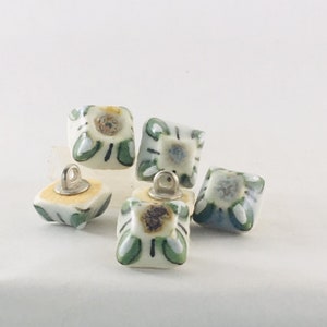 Op de afbeelding: Vijf vierkante keramische knopen met een witte achtergrond en groene, blauwe en gele bloemmotieven. Elke knop heeft een kleine zilveren lus aan de bovenkant.