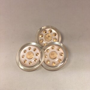 May include: Three clear, round buttons with a central gold-colored spiral design and surrounding gold-colored accents. The buttons have a slightly raised, transparent rim. These decorative buttons are suitable for sewing and craft projects.