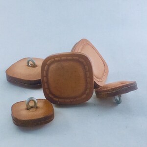 May include: Four brown leather square buttons with a stitched border and a metal loop on the top.