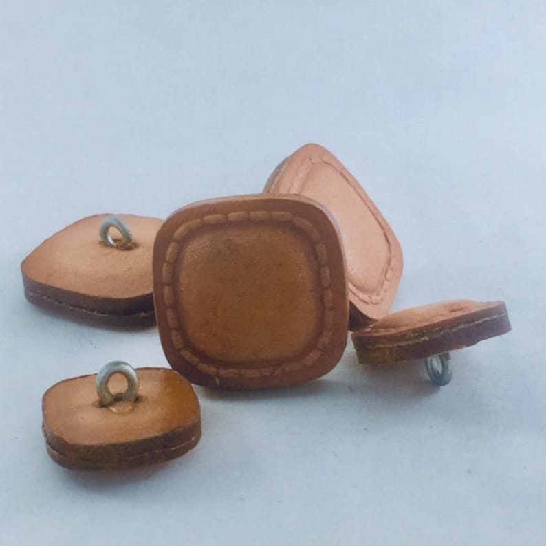 May include: Four brown leather square buttons with a stitched border and a metal loop on the top.