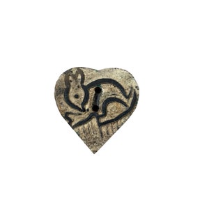 Puede incluir: Un botón de madera en forma de corazón con un diseño tallado de un conejo. El botón es de color marrón claro con detalles negros.