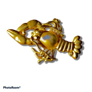 May include: A gold-colored lobster brooch with intricate details. The lobster is depicted with its claws raised and its tail curled.