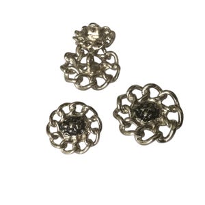 May include: Three silver-coloured buttons with a chain-link design. The buttons have a floral shape with a dark centre. One button has a loop on the back, suggesting it's a shank button. The buttons are on a white background.