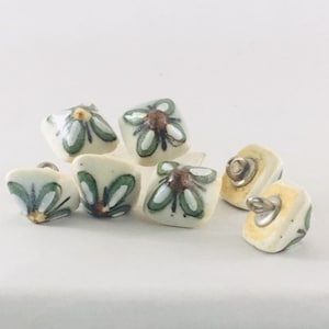 Puede incluir: Seis botones de cerámica con fondo blanco y diseños florales verdes y marrones. Algunos botones tienen forma cuadrada, mientras que otros tienen forma triangular. Algunos botones tienen un vástago de metal.
