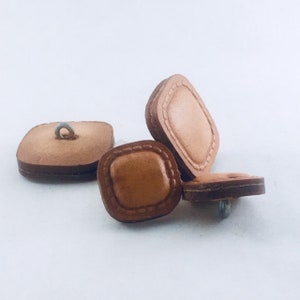 May include: Three brown leather buttons with a square shape and stitched edges. The buttons have a smooth surface and a hole for sewing.
