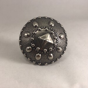 Puede incluir: Un pomo decorativo redondo plateado con una superficie texturizada y un diseño central cuadrado. El pomo está adornado con pequeños detalles circulares y puntiformes en relieve. Esta pieza de ferretería es adecuada para muebles o armarios. El pomo mide unos 5 cm de diámetro.