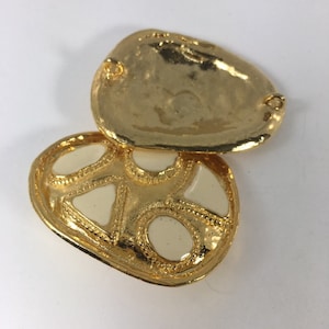 Może przedstawiać: Złote, ozdobne pudełko z zawiasem i wzorem z białej emalii. Pudełko ma kształt serca i fakturowaną powierzchnię.