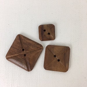 May include: Three wooden buttons in a square shape with rounded corners. The buttons are brown and have two holes each.