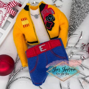 May include: A miniature yellow jacket with black and red accents, paired with a white shirt and blue jeans with a red belt. The jacket has embroidered details. A silver necklace is visible. The outfit is a Christmas ornament.