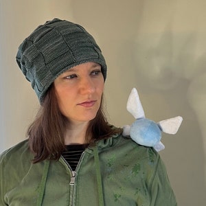 May include: A woman wearing a green hooded sweatshirt with a zipper and a dark green knit beanie. She is wearing a light blue plush toy on her shoulder.
