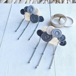 Peut inclure: Deux paires de boucles d'oreilles faites à la main avec un design en forme de nuage. Chaque boucle d'oreille présente un groupe d'éléments en pâte polymère en spirale grise, blanc cassé et gris foncé, suspendus à une chaîne argentée. Deux bagues en argent sont également visibles.