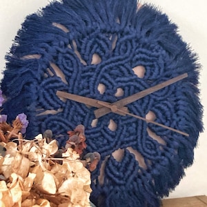 May include: A round, navy blue macrame wall clock with wooden clock hands. The clock has a fringed edge and a light-coloured backing. Dried flowers are visible in the lower left corner.