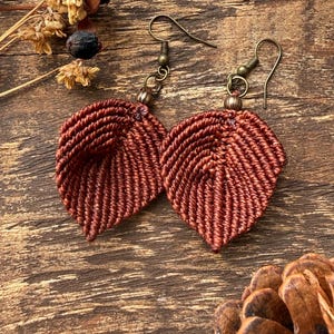 May include: A pair of rust-colored macrame earrings shaped like leaves. Each earring features a detailed cord-woven texture, a small metallic bead at the top, and a dark bronze-toned metal hook for attachment, showcasing a distinct fiber art design.
