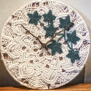 May include: A round macrame clock with a cream-coloured woven design. The clock features dark green leaf accents and wooden clock hands. The clock is a decorative wall hanging.