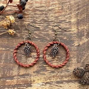 May include: A pair of dangle earrings featuring a circular hoop made of twisted rust-colored cord. Each hoop suspends a small, metallic bronze-toned pinecone charm from a circular ring attached to a bronze-toned fishhook ear wire.