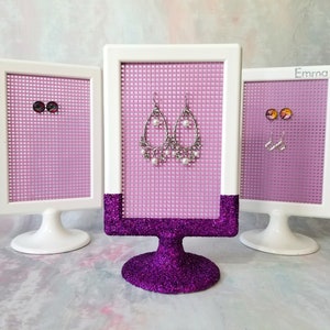 May include: Three white framed earring display stands with pink mesh backing. The center stand has a purple glitter base and displays a pair of silver dangle earrings with white pearls. The stands on either side have white bases and display pairs of stud earrings.