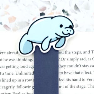 May include: A magnetic bookmark featuring a light blue cartoon manatee illustration attached to a solid dark blue rectangular base, positioned on an open book page with the text JustAddJewelry displayed in a white speech bubble shape below it.